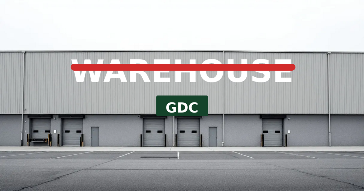 A plain gray commercial warehouse with the word WAREHOUSE in large white letters struck through in red, and a dark green GDC banner hanging below it